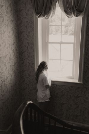 Black and white photo of a young girl in white dress standing by a vintage window with draped curtains.