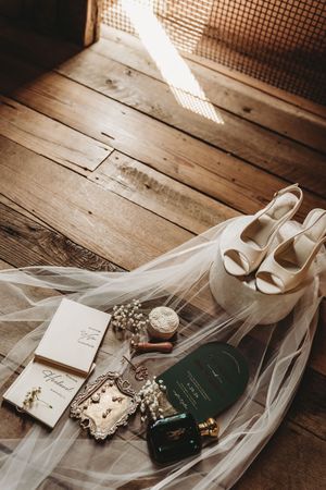Bridal flat lay with white heels, veil, wedding stationery, perfume, and jewelry on rustic wood floor.