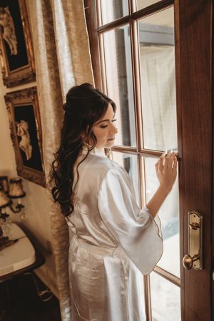 Bride in white satin robe standing by wooden window, looking down with elegant curly hair in vintage room.