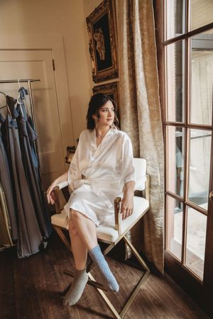 Bride in white satin robe and blue socks sits in gold chair by window with bridesmaid dresses on rack nearby.