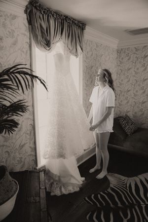 Bride in white pajamas admiring her wedding dress hanging by a sunlit window in a vintage room.