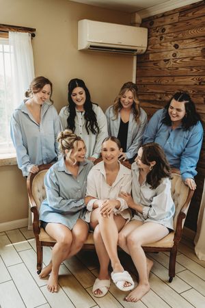 Bride and bridesmaids in blue shirts laughing together on a vintage sofa during wedding morning preparations.