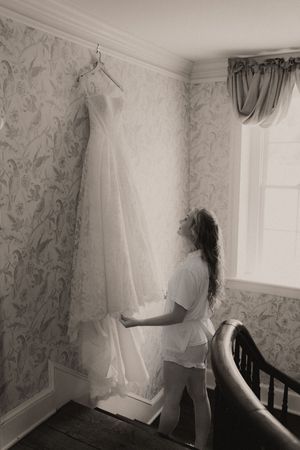 Black and white photo of a bride admiring her wedding dress hanging on a wall in an elegant room.