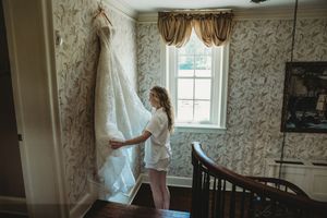 Bride in white robe admiring her wedding dress hanging on wall in elegant floral wallpapered room.