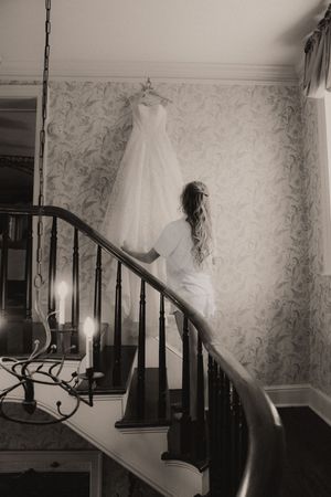 Bride in robe admiring wedding dress hanging on staircase in elegant black and white photo.