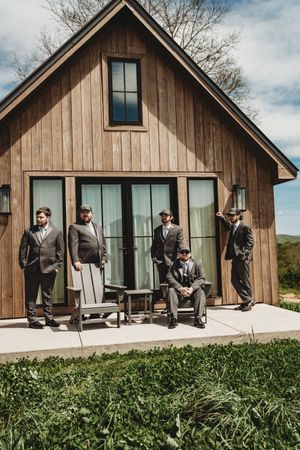 Four groomsmen in suits pose outside a rustic wooden barn venue on a sunny wedding day.