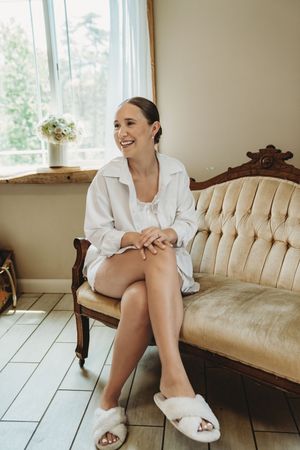 Smiling bride in white robe and fluffy slippers sitting on vintage tufted sofa near window on wedding morning.
