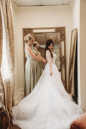Bride in white lace ballgown wedding dress being helped by bridesmaid in sage green dress before large ornate gold mirror.