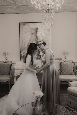 Black and white photo of a bride and bridesmaid sharing a tender moment in an elegant room with chandelier.