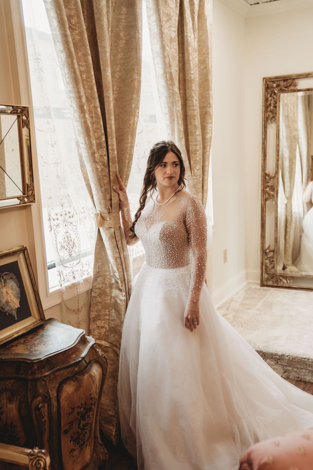 Bride in beaded long-sleeve wedding gown stands by draped window in elegant room with ornate mirror.