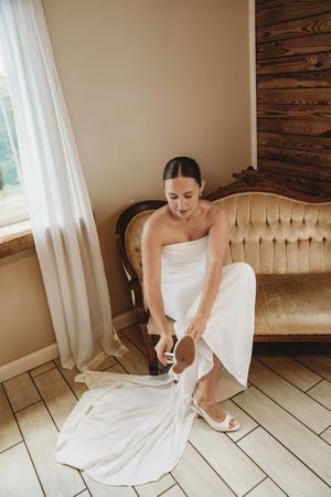 Bride in white strapless wedding gown sitting on vintage gold sofa putting on white heels before ceremony.