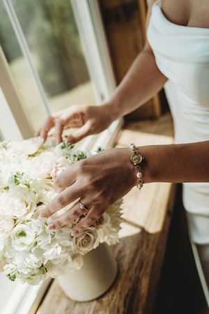 Bride arranging white rose bouquet in vase by window, wearing elegant bracelet and ring on wedding day.
