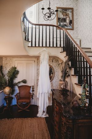 White wedding dress hanging on curved staircase in elegant vintage home with ornate wallpaper and antique furnishings.