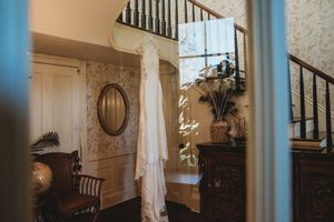 Elegant wedding dress hanging on a staircase in a vintage-styled home with ornate wallpaper and antique furniture.