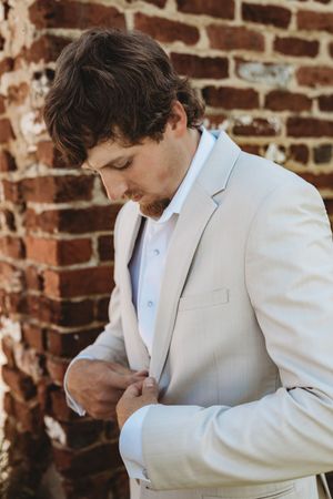A groom in a beige suit adjusts his jacket button against a rustic brick wall backdrop.
