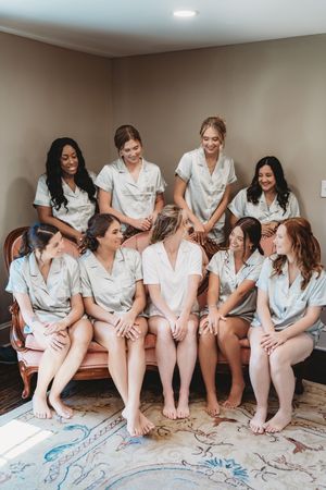 Bridesmaids in matching sage green pajamas gather together smiling, with bride in white, during wedding morning preparations.