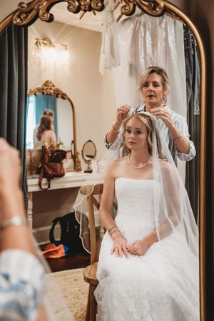 Mother helping bride with veil in ornate gold mirror reflection during wedding preparations.