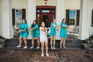 Bridesmaids in teal robes celebrate with the bride in white outside a white Southern mansion.