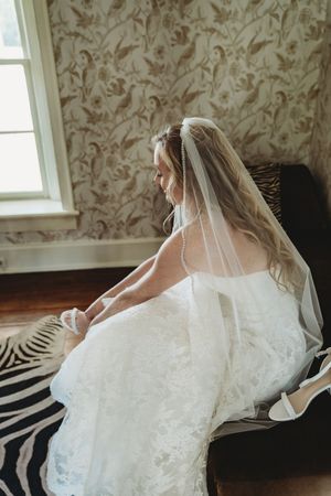 Bride in lace wedding gown and veil sits by window on zebra rug, adjusting her shoe in elegant floral wallpaper room.