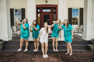 Bridesmaids in teal robes pose playfully on white venue steps with bride in white pajamas.