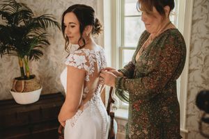 A bride in a lace wedding gown has her dress fastened by a woman in a floral dress in an elegant room.