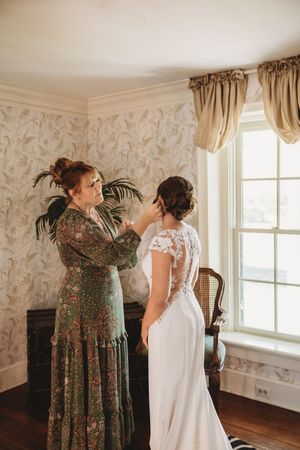 A woman in a floral dress helps a bride in a lace wedding gown prepare near a sunlit window.