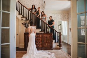Bride in elegant white gown reveals look to bridesmaids on staircase in vintage home interior.