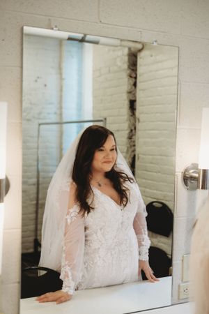 Bride in lace dress posing elegantly against white brick wall with natural lighting.