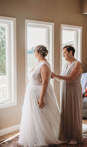 Two people in elegant dresses preparing for a wedding by sunlit windows.