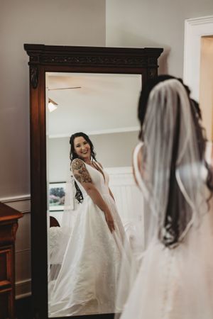 Bride in white dress admiring her reflection in vintage wooden mirror.