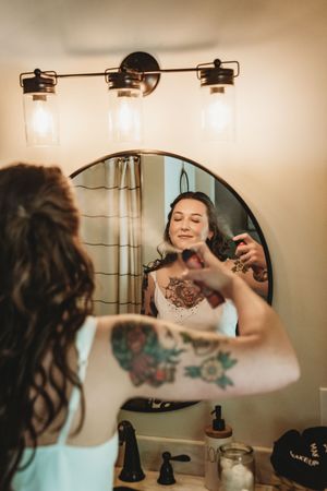 Tattooed woman spraying setting spray on her makeup while looking in a round bathroom mirror.