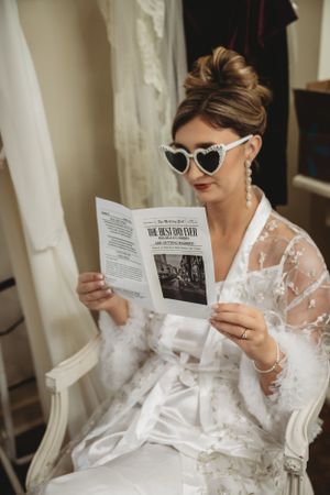 Bride wearing sunglasses and lace robe reads a newspaper while getting ready on her wedding day.