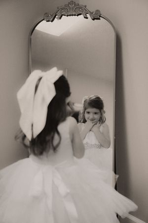 Black and white photo of a young girl in a white dress admiring herself in an ornate vintage floor mirror.