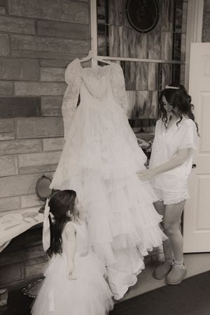 Black and white photo of a bride and flower girl admiring a lace wedding gown hanging in a doorway.