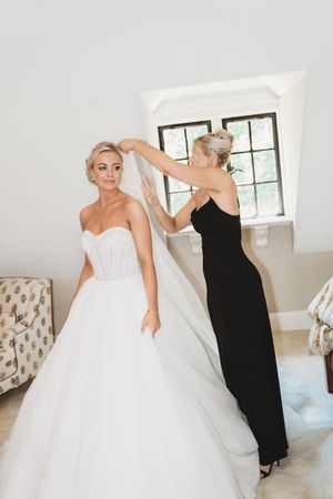 A bride in a voluminous white wedding gown gets help with final dress adjustments from someone in black.