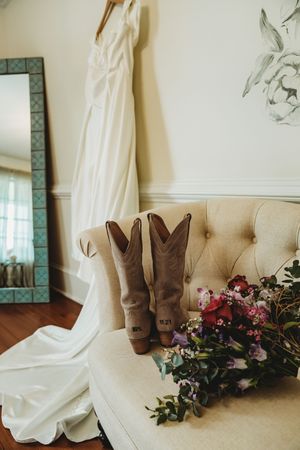Bridal flat lay with cowboy boots, white wedding dress, and colorful floral bouquet on a tufted chair.