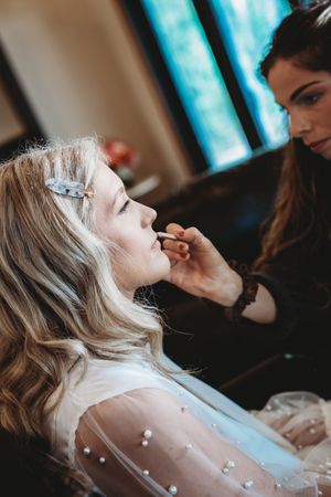 Makeup artist applying lipstick to a blonde bride wearing a pearl-embellished white gown before her wedding.