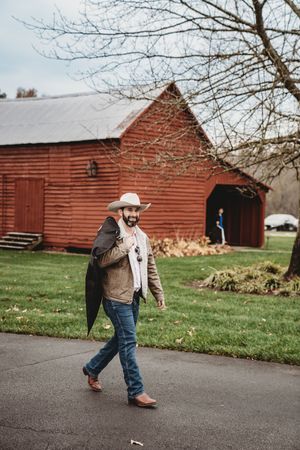 Man in cowboy hat and jeans walking on driveway near red barn on cloudy day.