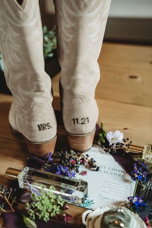 Bridal cowboy boots with MRS. and 11.21 on heels, surrounded by wedding invitation and wildflowers.