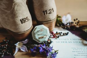 Wedding flat lay featuring MRS. monogrammed shoes dated 11.21, diamond ring in velvet box, gold bands, and floral invitation.