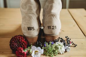 Cowboy boots stamped with MRS. and 11.21 wedding date surrounded by colorful wildflowers on a wooden table.