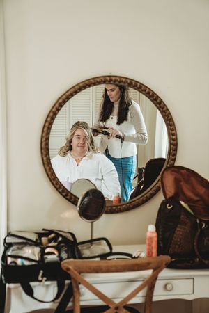 A hairstylist curling a woman's hair reflected in a round ornate mirror above a vanity table.