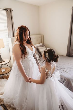 A bride in a white lace gown holds hands with a flower girl in a matching dress in a softly lit bedroom.