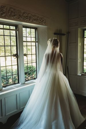 Elegant white wedding dress with flowing tulle skirt and veil displayed in a sunlit room with ornate windows.