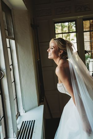 A bride in a strapless white gown and veil poses by a window in natural light.