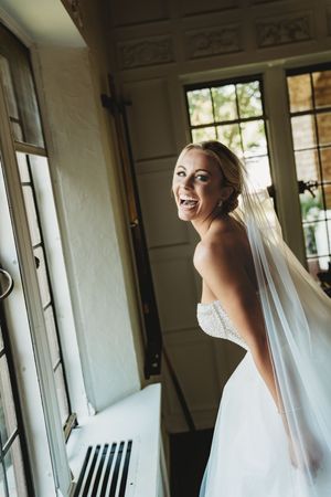 Bride in white wedding dress and veil poses by window in elegant interior setting.