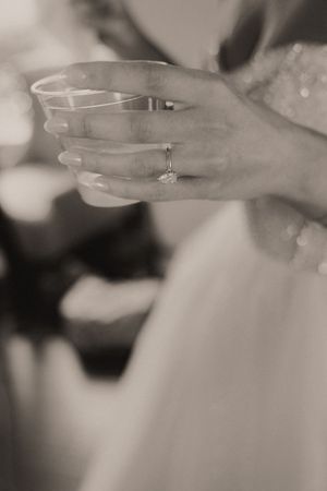 Close-up black and white photo of hands holding a glass, showing a wedding ring and lace dress details.