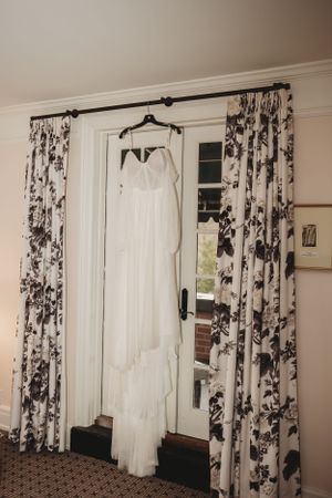 White wedding dress hanging against black and white floral curtains in preparation room.