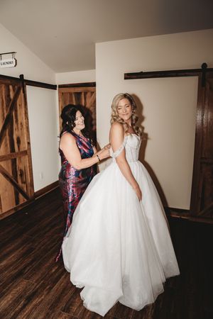 A bride in a white ballgown getting help with her dress from a woman in a floral outfit in a rustic barn venue.