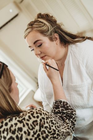 A makeup artist applying lip liner to a client wearing a leopard print top in a bright room.